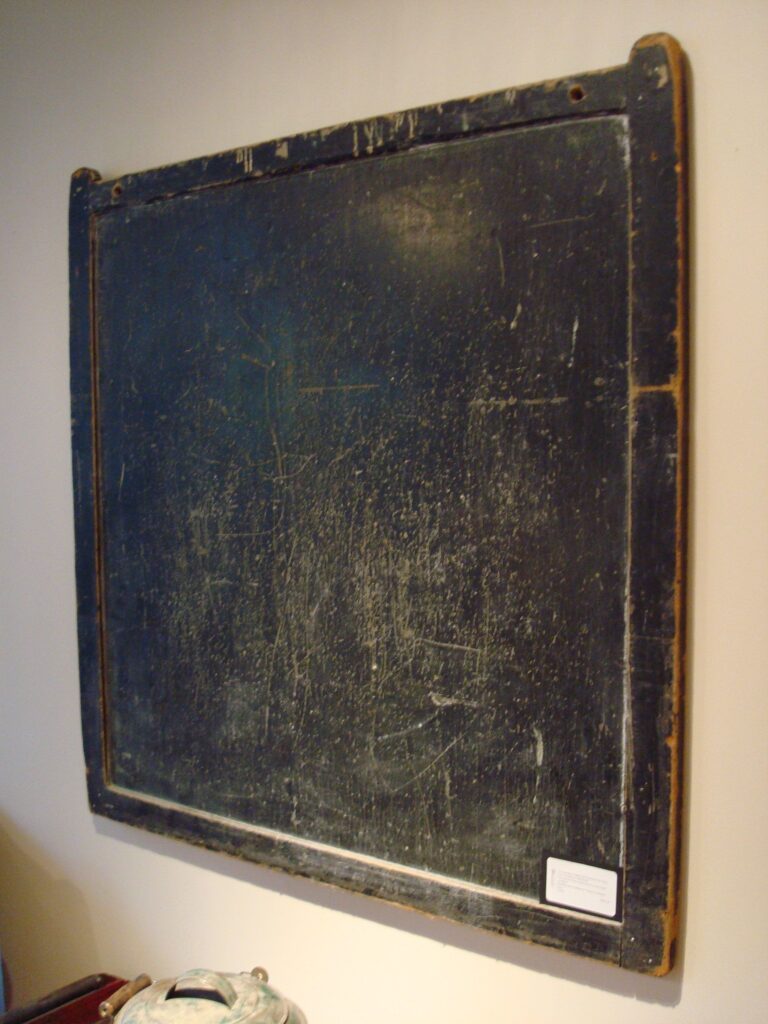 An old black chalkboard hangs on a white wall in a classroom.
