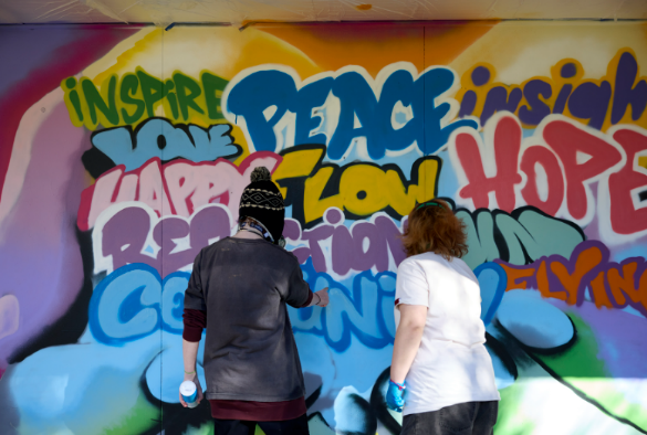 Two women spray painting graffiti on a wall. Some of the words they are painting include, "Peace", "Inspire","Love", "Hope", "Happy".