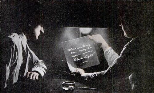 Two men looking at a piece of paper which is help under light to reveal the secret message.   