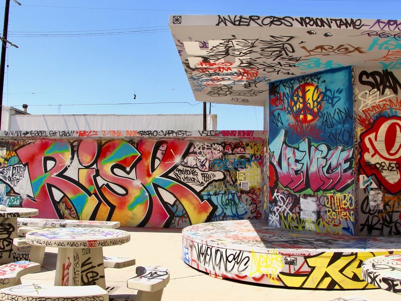Graffiti-covered benches and tables in a vibrant skateboard park, showcasing urban art and adding a unique touch to the surroundings. The main graffiti painting has the word "risk" written in colorful painting.